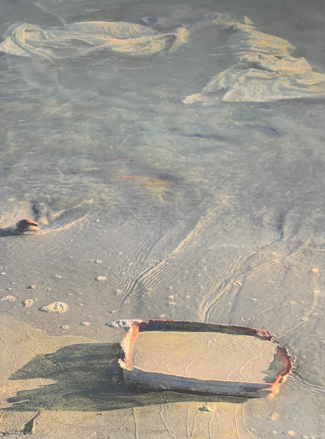 Segundo premio del Concurso de pintura de BBVA, Título: Contaminación, esta pintura muestra una escena de una playa en primer plano, donde la arena húmeda queda marcada por suaves ondulaciones y texturas creadas por el agua al retirarse. La luz es cálida y baja, proyectando sombras alargadas, en primer plano donde se junta la arena con el mar hay un recipiente metálico ya oxidado parcialmente hundido en la arena.. 